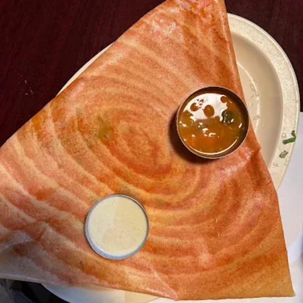 A large, folded dosa with two small bowls of chutney and sambar on a white plate.