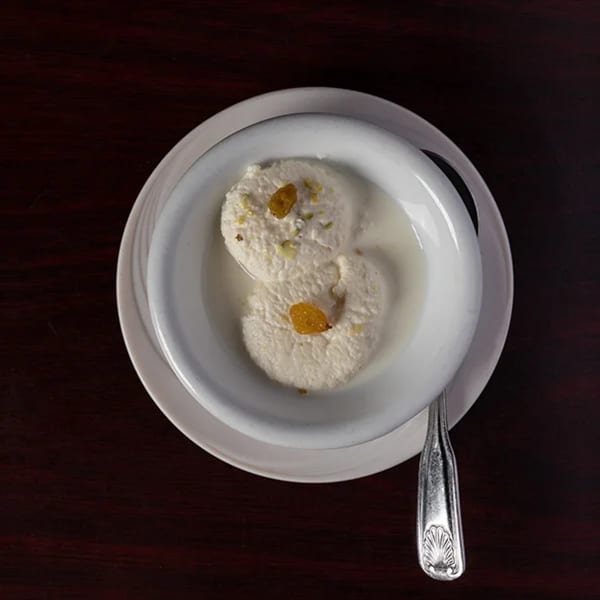 Two rasmalai pieces in a white bowl with a silver spoon on a dark wooden table.